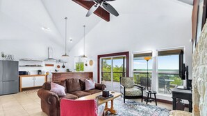 A spacious living area with a brown leather sofa, a coffee table, and a red chair. A kitchen is visible in the background with a white ceiling and a ceiling fan. - Canyon Breeze I 3 Bedroom I Pet Friendly I Canyon Lake! (Canyon Lake)
