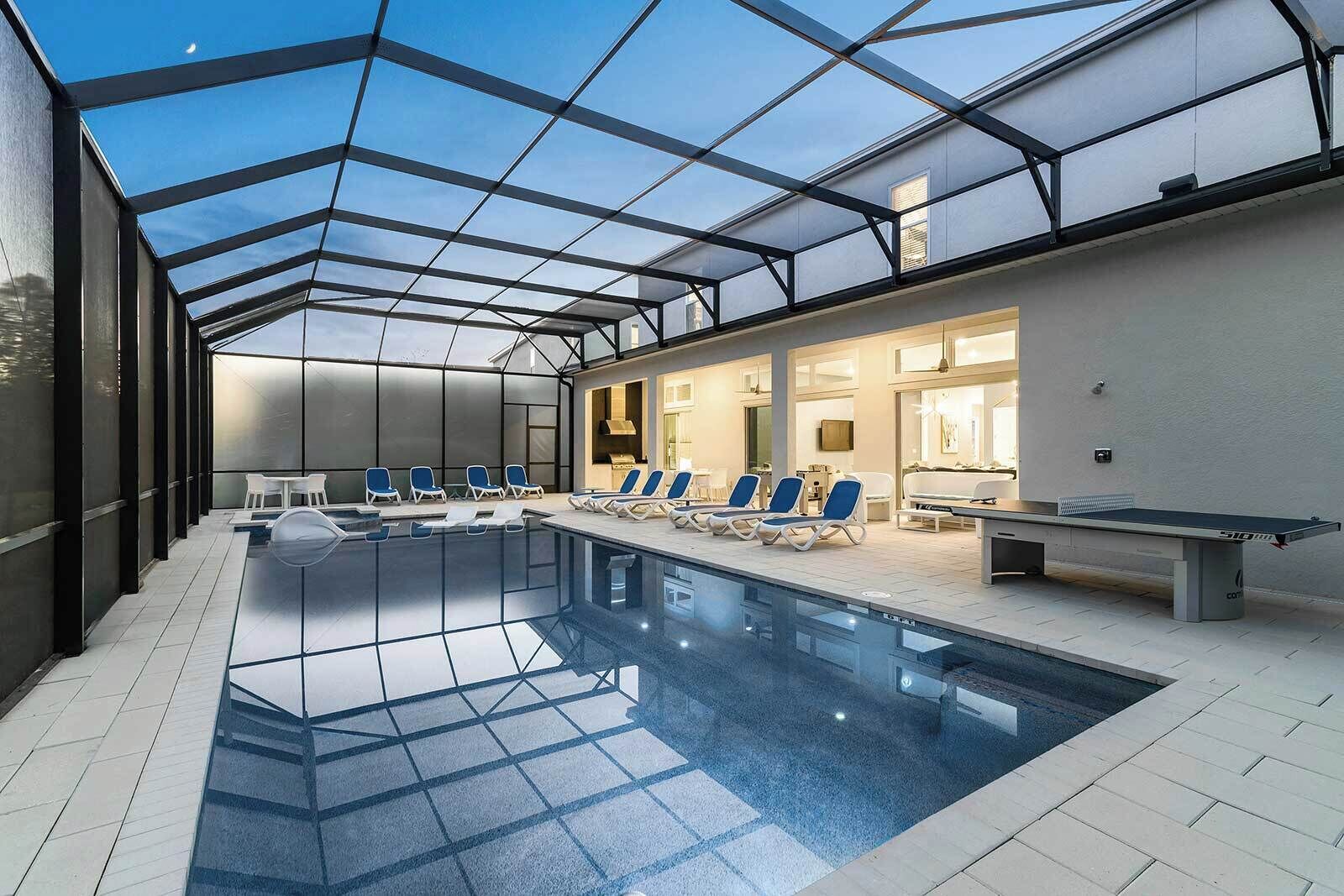 An indoor swimming pool with clear glass ceilings, lounge chairs, and a table with a ping pong table.