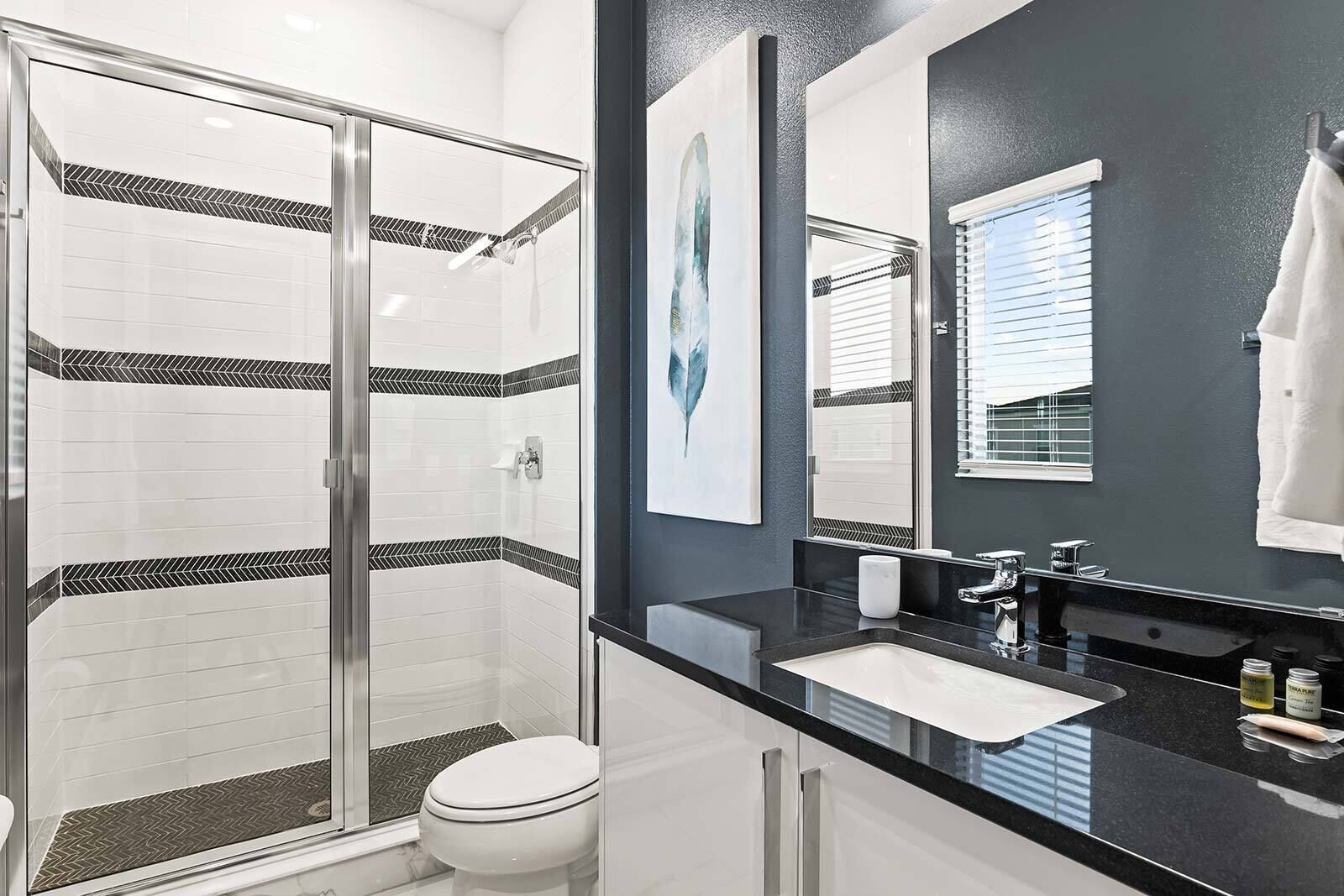 Un baño moderno con cabina de ducha de vidrio, inodoro blanco, encimera negra con lavamanos y un espejo grande.