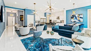 A modern living room with a blue sofa, white armchairs, and a dining area with a blue table. - Fun Home w/ Trampoline RM, Pirate Theme BR, Arcade, Theater & Gym - Near Disney (Davenport)