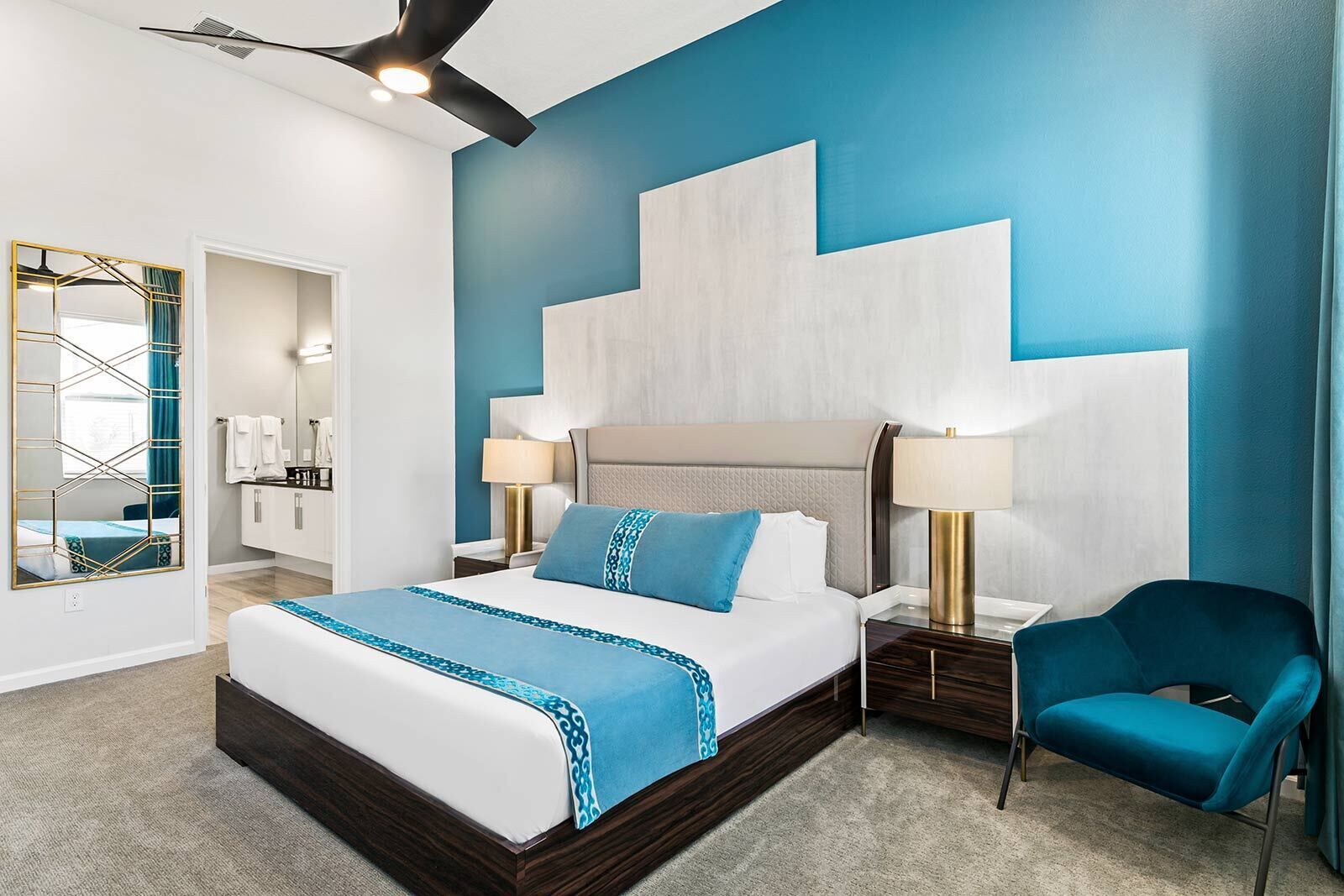 Ein modernes Hotelzimmer mit einem großen Bett, zwei Nachttischen mit Lampen, einem blauen Sessel und einem Spiegel mit geometrischem Design.