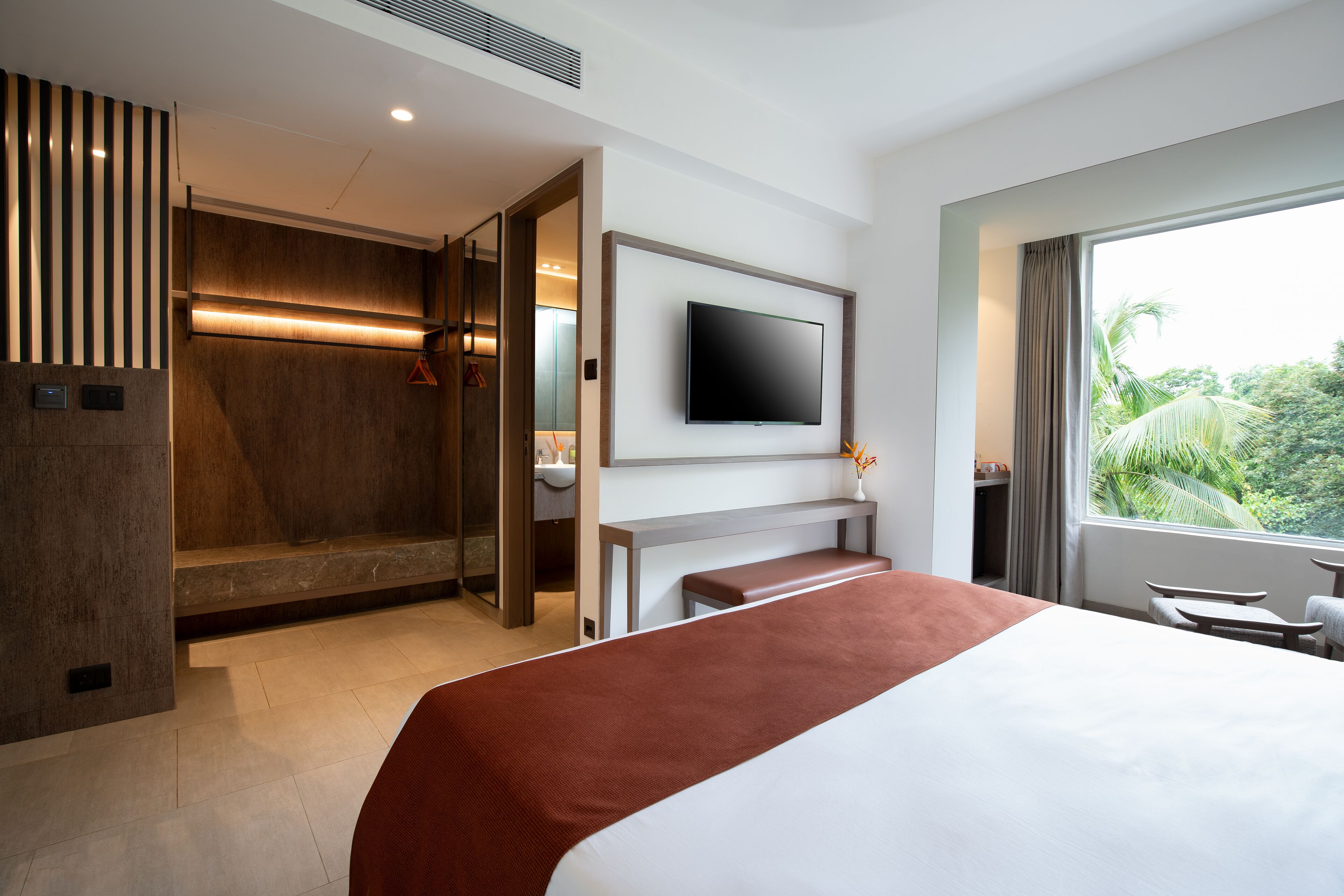 A modern hotel room with a large bed, a flat-screen TV, a bench, and a window with a view of greenery.