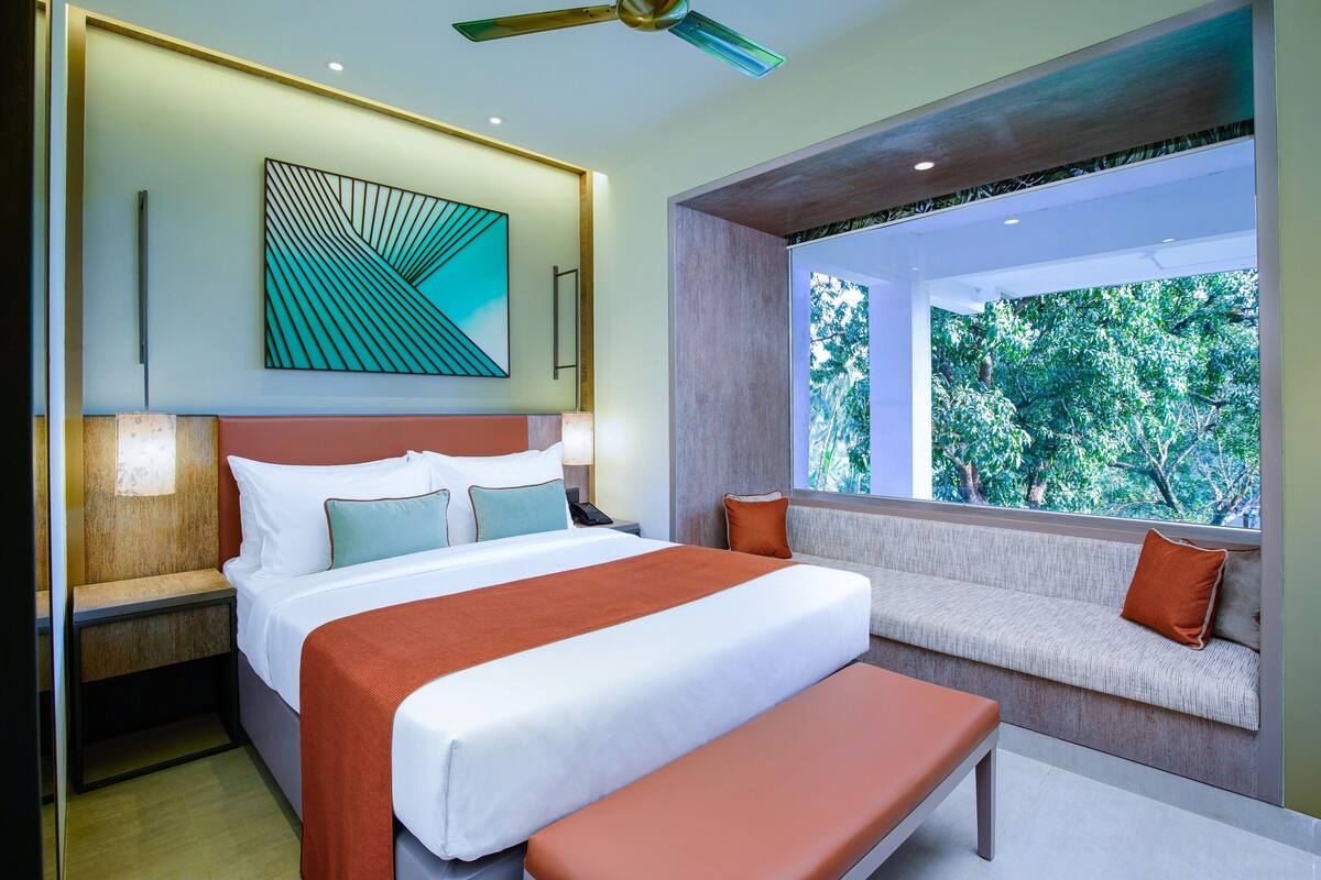 A modern hotel room with a large bed, a bench, a sofa, and a window with a view of trees.