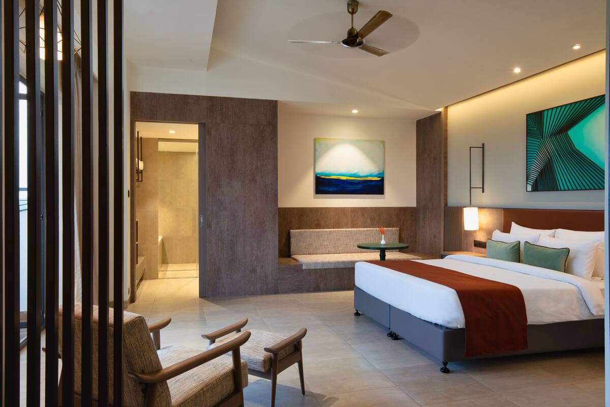 A modern hotel room with a large bed, a seating area with chairs, a painting on the wall, and a ceiling fan.