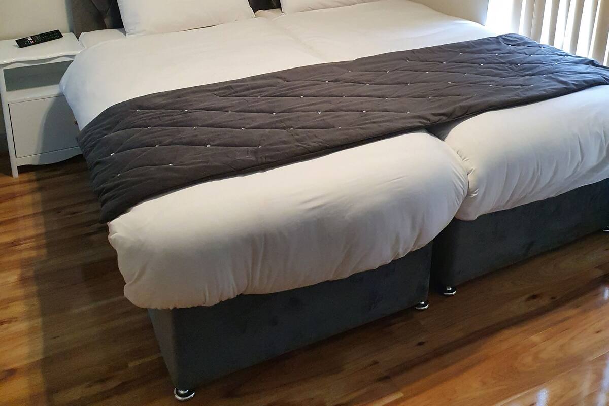 A double bed with a dark headboard and footboard, white bedding, and a dark quilt. There is a bedside table with a remote control on it. The room has wooden flooring and a window with blinds.