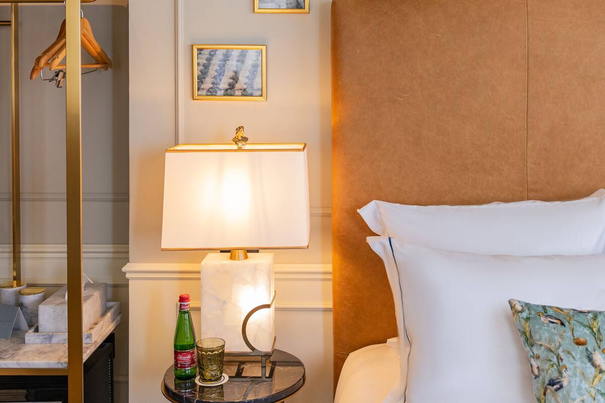 A hotel room with a bed, a bedside table with a lamp, a small table with a bottle and glasses, a coat rack, and two framed pictures on the wall.