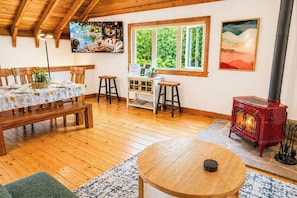 Une pièce chaleureuse avec un plancher en bois, une table à manger recouverte d’une nappe, un banc, une petite table ronde en bois, un poêle à bois rouge, une fenêtre donnant sur la verdure et un tableau accroché au mur.
