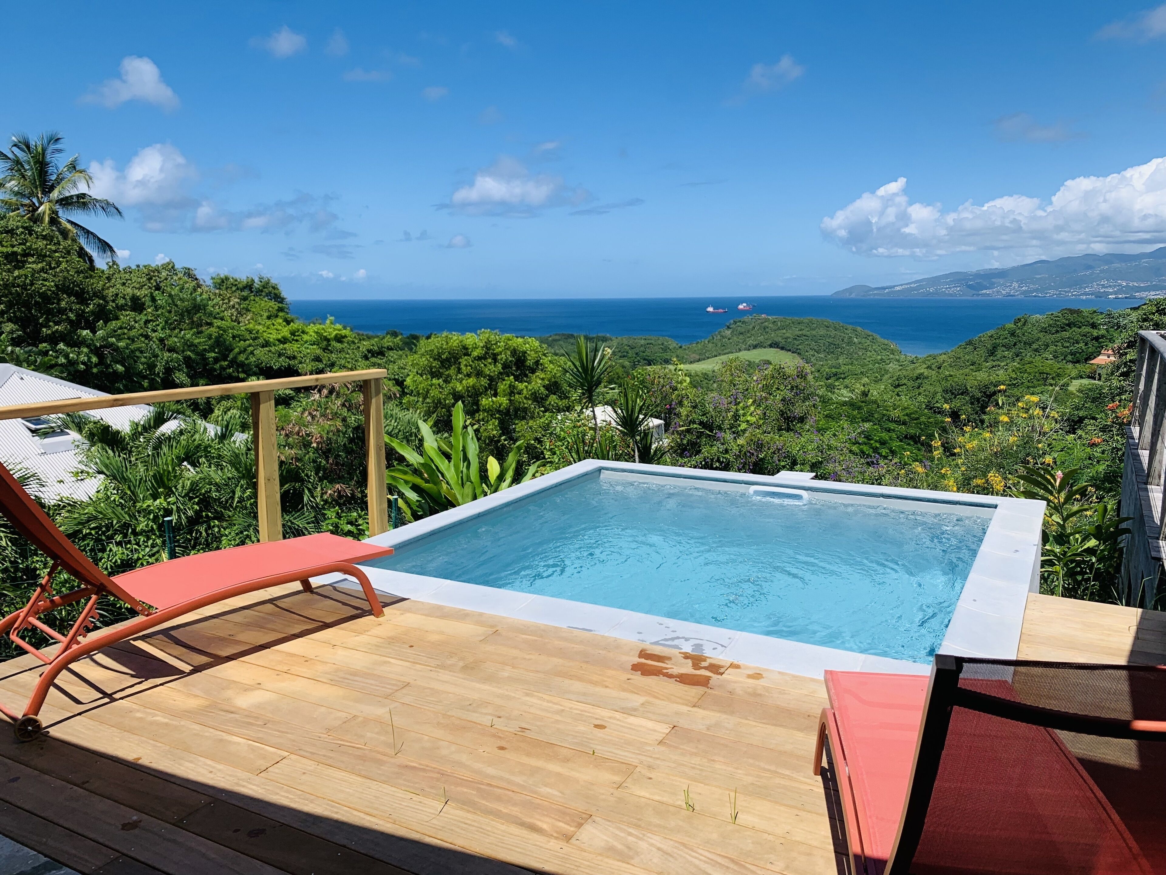 A pool with a wooden deck, red lounge chairs, and a view of the ocean and hills.
