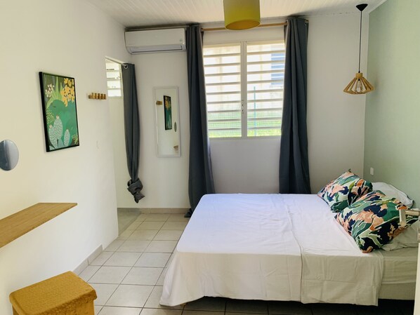 A bedroom with a bed, a bedside table, a mirror, a window with curtains, and an air conditioning unit. - Villa Frisch & Gemütlich, Trois-Ilets, Swimming Pool, Ocean View, 3***, France (Les Trois-Ilets)