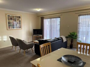 A living room with a sofa, two armchairs, a dining table, a TV, and a framed collage on the wall.