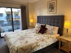 A hotel room with a large bed, two bedside lamps, a window with a view of buildings, and a framed picture on the wall.