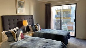 A hotel room with two beds, a nightstand, a lamp, a framed picture on the wall, and a balcony with a chair.