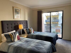 A hotel room with two beds, a nightstand, a lamp, a framed picture on the wall, and a balcony with a chair.