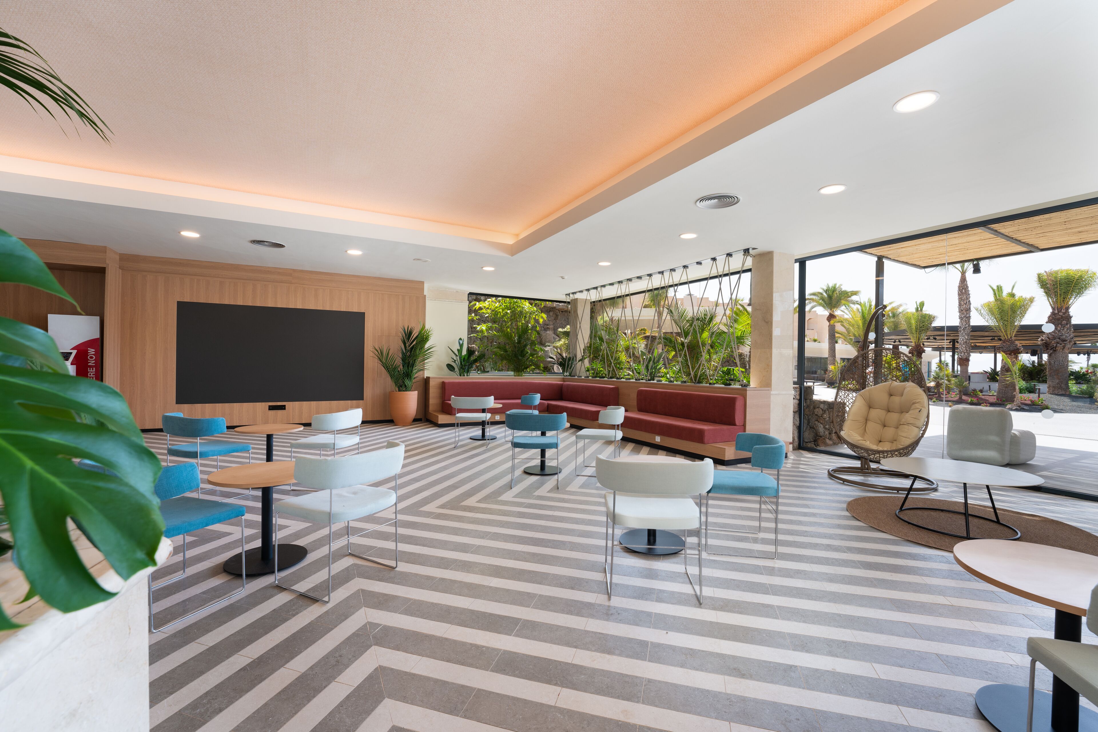 A modern indoor lounge area with striped flooring, multiple seating options including sofas and chairs, a large flat-screen TV, and a view of greenery outside.