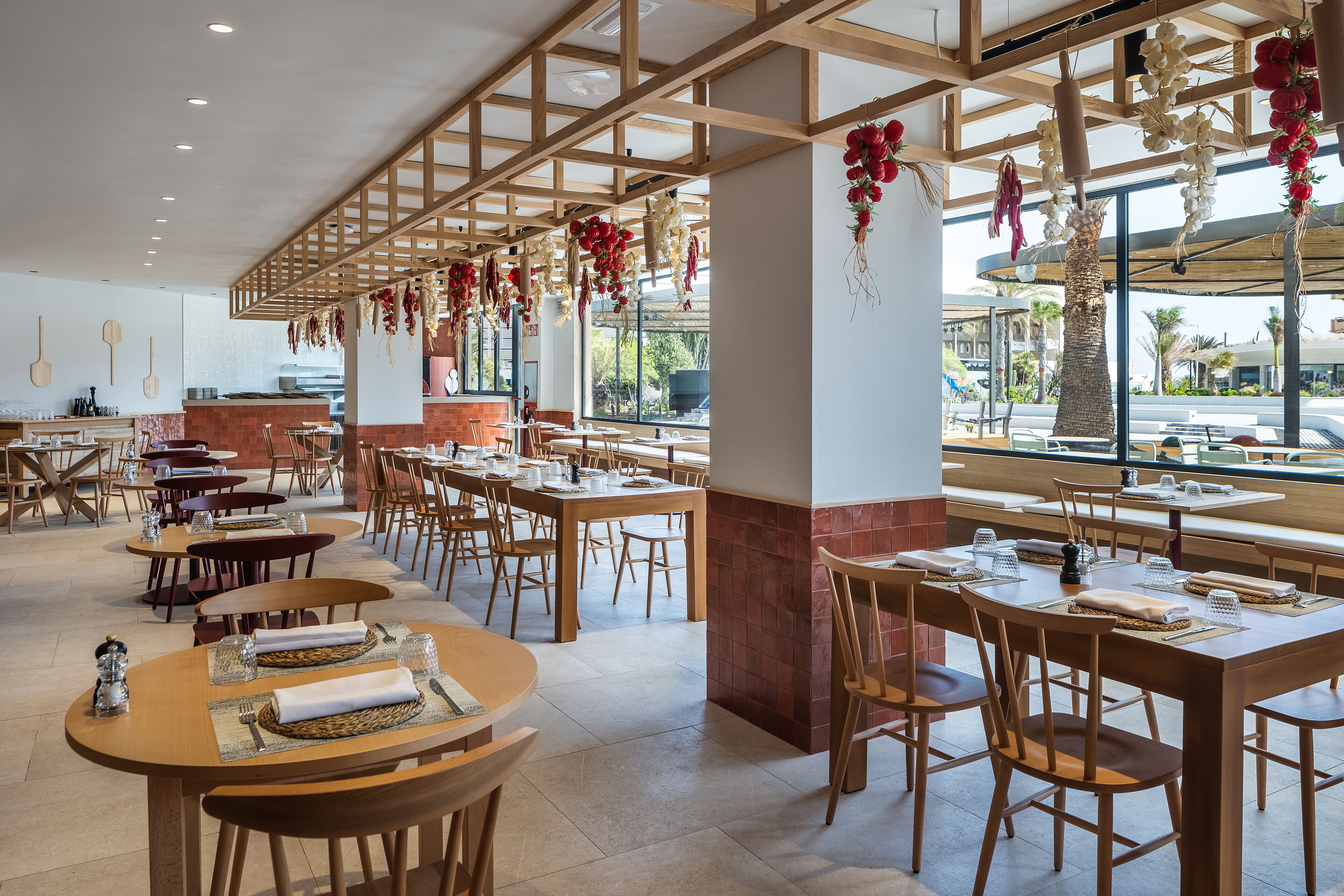 Un restaurant spacieux, doté de nombreuses tables et chaises, de poutres en bois au plafond et de grandes fenêtres offrant une vue sur l’extérieur.