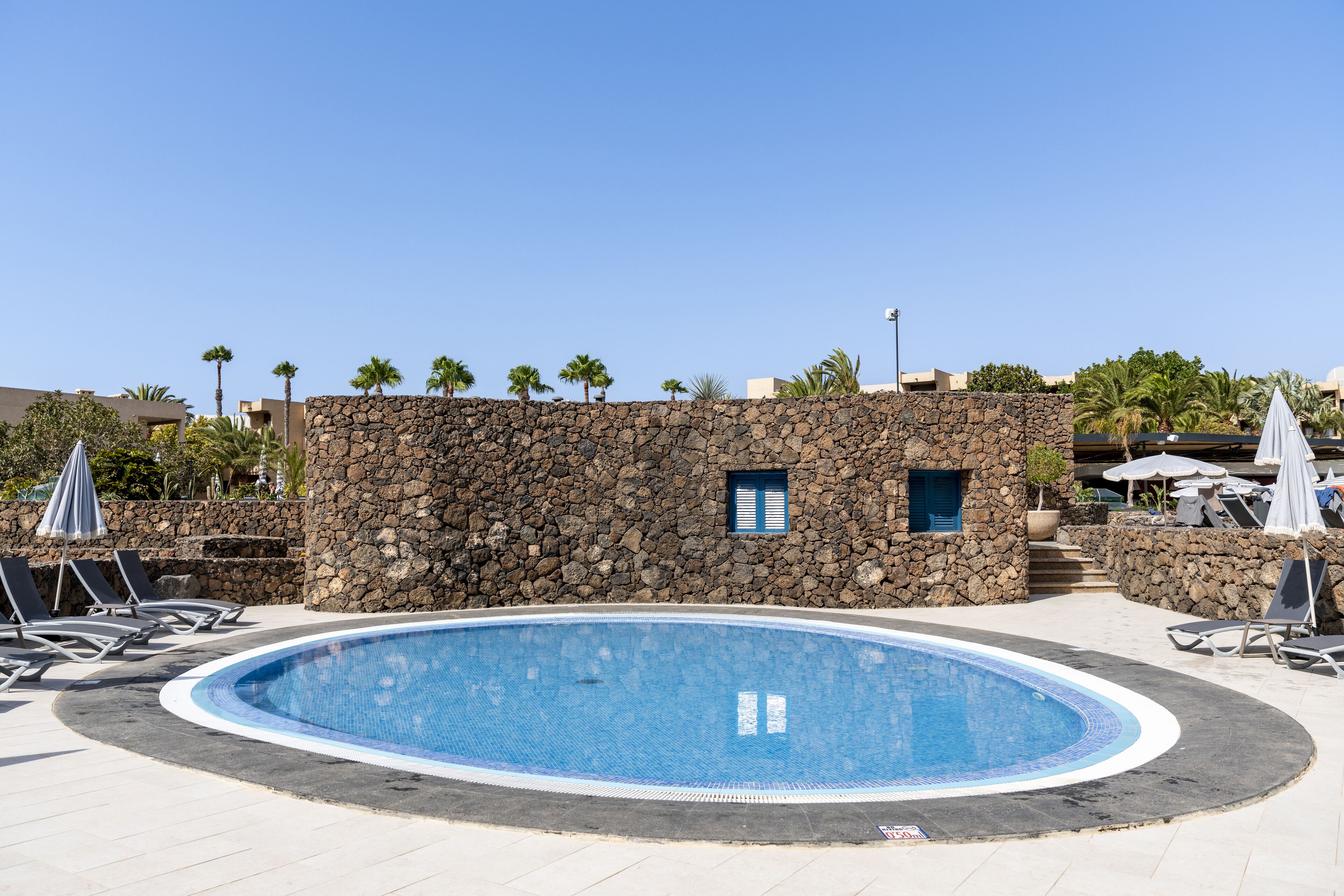 Una piscina rodeada de sillones de descanso y sombrillas, con un muro de piedra y palmeras al fondo.