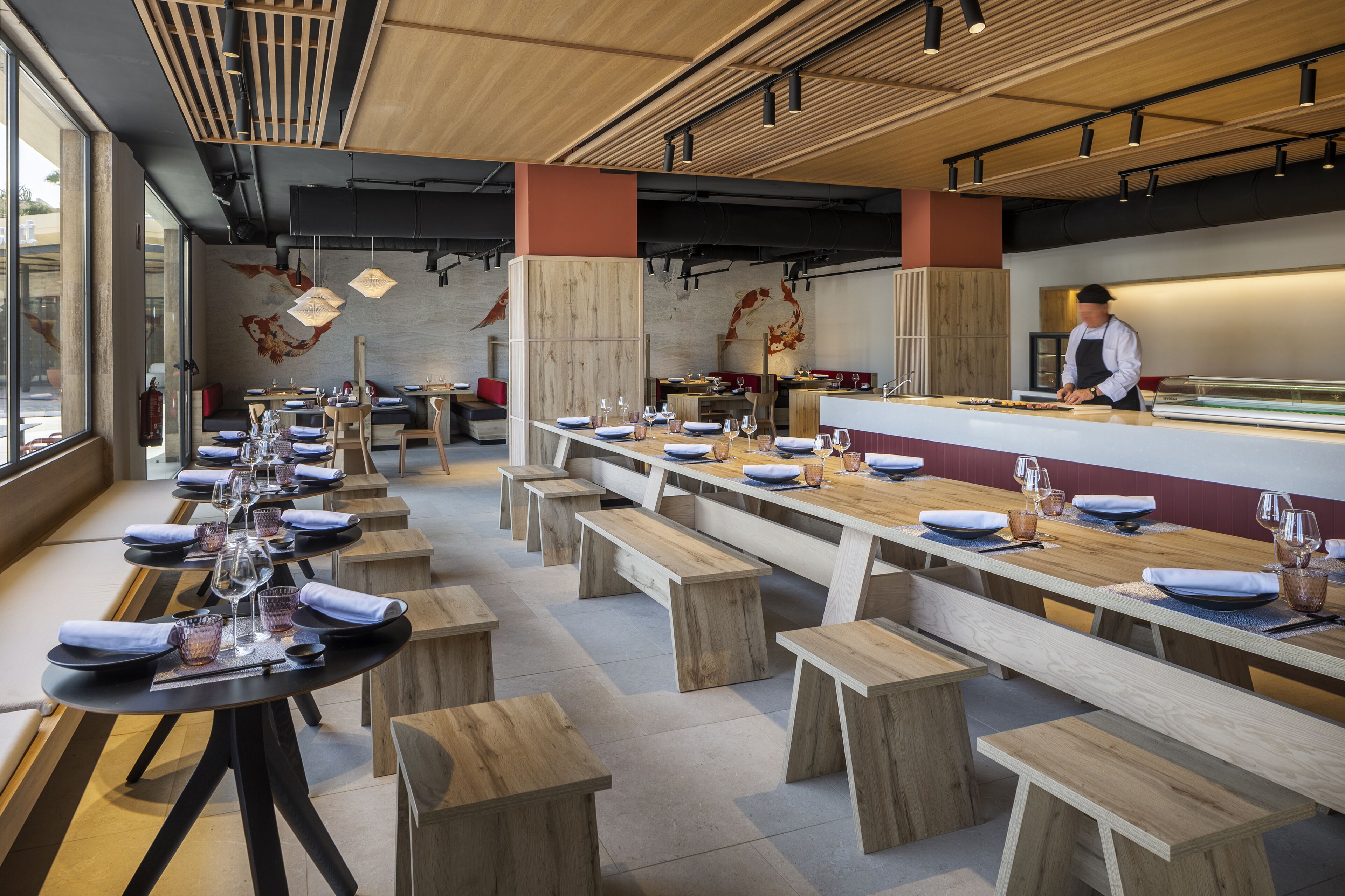 Un restaurant moderne avec des tables et des bancs en bois, un chef de cuisine derrière le comptoir et une grande fenêtre sur le côté gauche.