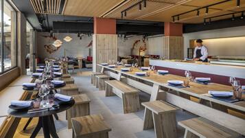 Un restaurant moderne avec des tables et des bancs en bois, un chef de cuisine derrière le comptoir et une grande fenêtre sur le côté gauche.