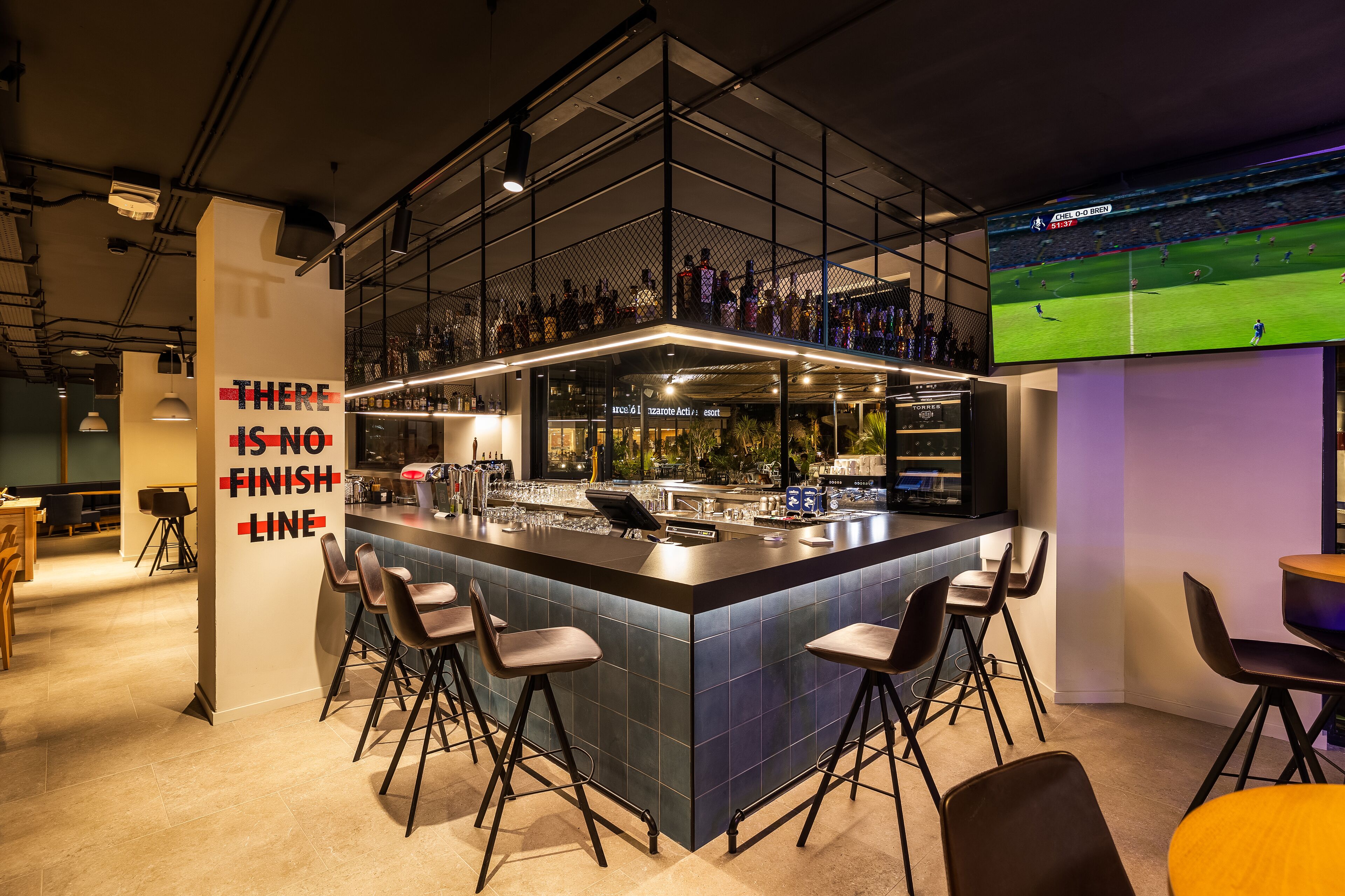 A modern bar area with a large screen displaying a sports game, bar stools, and a sign stating "THERE IS NO FINISH LINE".