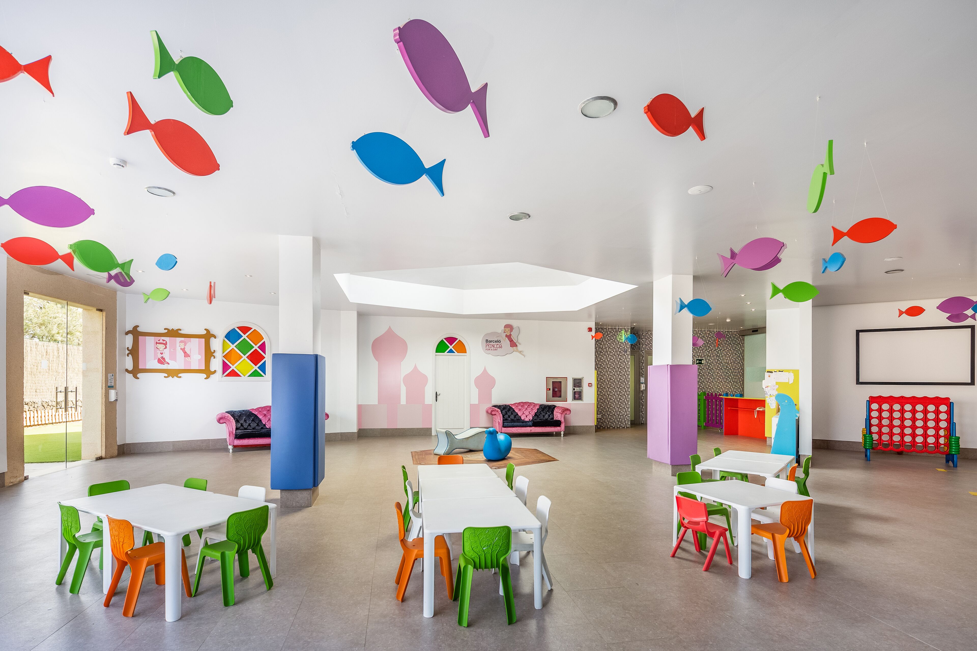 A brightly colored children's playroom with tables and chairs, fish decorations on the ceiling, and a large window.