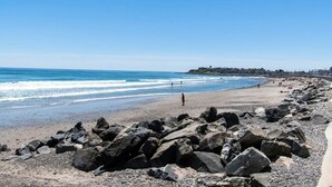 A rocky beach with waves, a clear sky, and a distant town. - Beachfront Getaway-06 (Hampton)