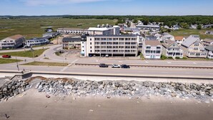 A coastal town with a large building with multiple balconies, a road with cars, and a rocky shoreline. - Beachfront Getaway-06 (Hampton)
