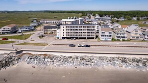 A coastal town with a large building with multiple balconies, a road with cars, and a rocky shoreline. - Beachfront Getaway-06 (Hampton)