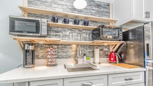 A modern kitchen with a microwave, toaster oven, and coffee maker. There are shelves above the sink with cups and a kettle. - Beachfront Getaway-06 (Hampton)