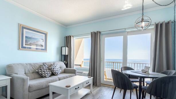 A living room with a sofa, coffee table, and dining table with chairs. There is a large window with curtains, a framed picture on the wall, and a hanging light fixture. - Beachfront Getaway-06 (Hampton)