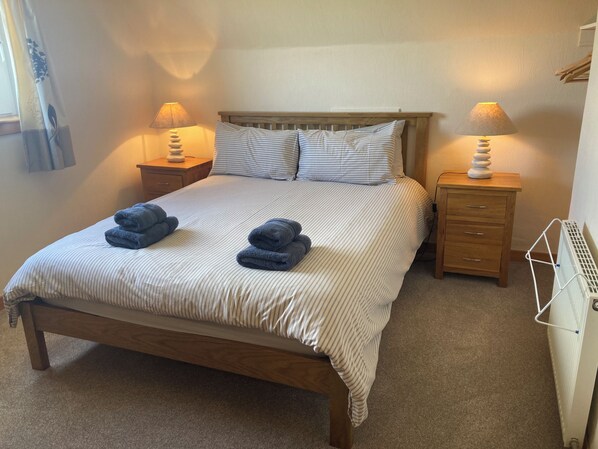 A neatly made bed with striped bedding and two folded towels. There are two bedside tables with lamps, a wooden headboard, and a radiator.