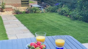 Un tavolo con una ciotola di fragole e due bicchieri di succo d'arancia, su un patio con vista su un giardino e una casa.