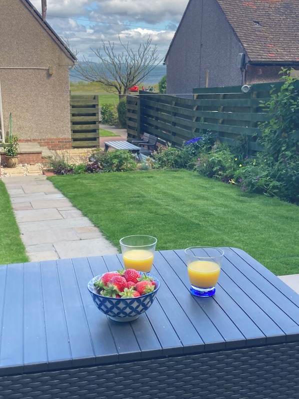 A table with a bowl of strawberries and two glasses of orange juice, set on a patio with a view of a backyard and a house. - Beach Cottage Sleeps 4 (2 Bedroom) (Sandhead)