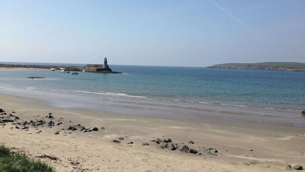 Paesaggio costiero con un faro, mare calmo e una spiaggia sabbiosa con rocce sparse.