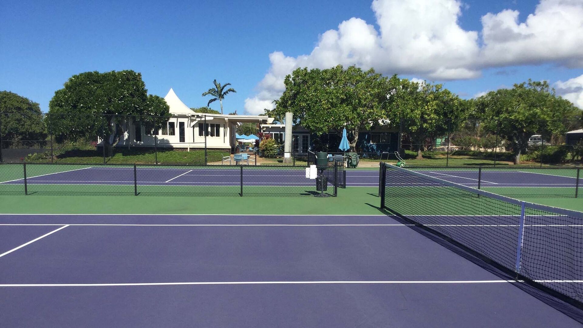 Una cancha de tenis al aire libre con red, rodeada de árboles y un edificio de fachada blanca.