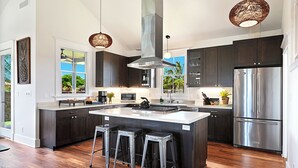 A modern kitchen with dark cabinetry, a large island with stools, stainless steel appliances, and pendant lighting. - Plantation House by Destination Kauai - Poipu Beach Estates, Central A/C, 3BD (Koloa)