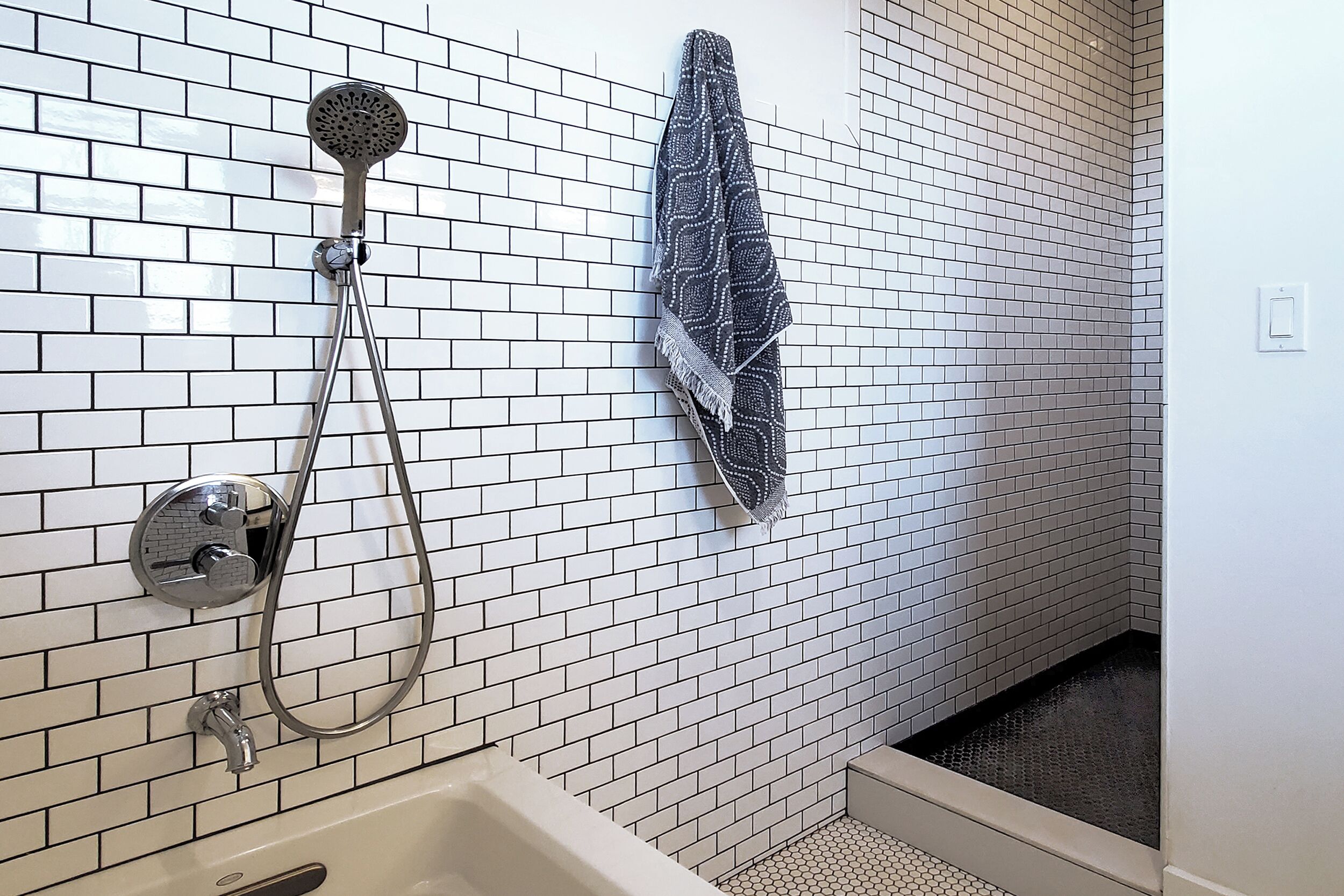 A bathroom with white tiled walls, a shower with a handheld showerhead, and a towel hanging on the wall.