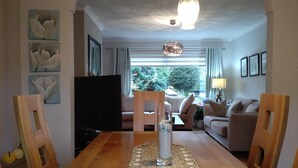 A cozy living room with a wooden dining table, a television, and a view of a garden through large windows.