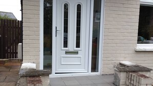 A white front door with a small step leading up to it, flanked by two stone walls and a window to the right.