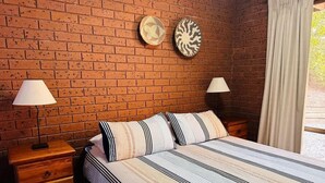 Un dormitorio con pared de ladrillo, una cama con ropa de cama a rayas, dos mesitas de noche con lámparas y dos relojes de pared.