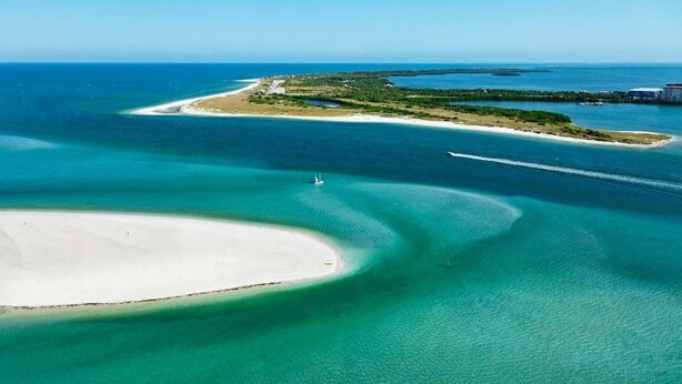 Vue aérienne d’une zone côtière avec des eaux turquoise limpides, une plage de sable et une petite île abritant des bâtiments.