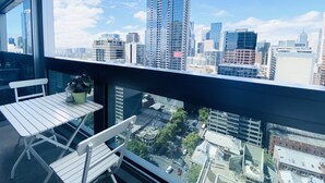 Un balcón con una silla y una mesa blancos, con vistas a un paisaje urbano de edificios modernos y cielo despejado.