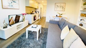 Una sala moderna con un sofá gris, una mesa de centro y un televisor. Hay una zona de cocina con armarios blancos y una mesa de comedor con sillas.