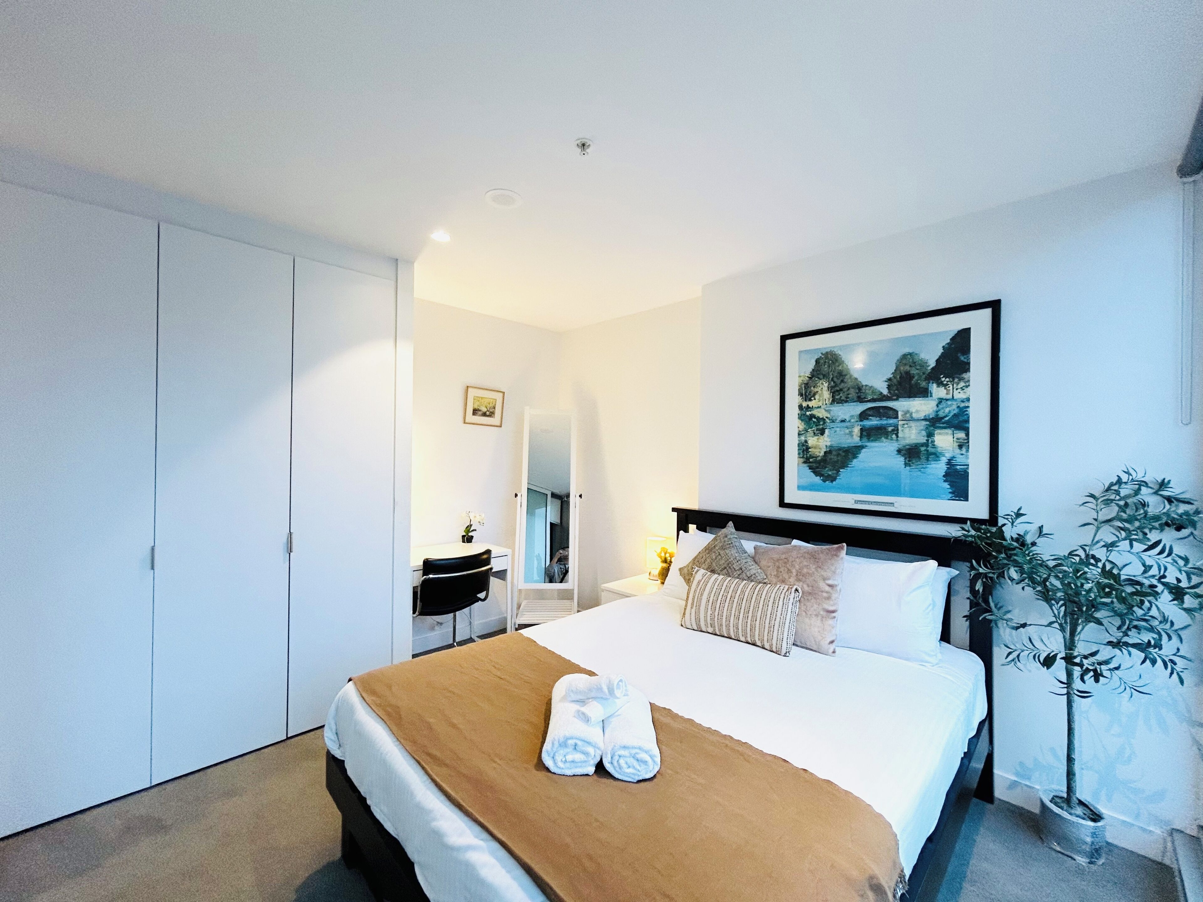 A hotel room with a bed, a desk, a chair, a potted plant, and a framed picture on the wall.