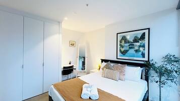 A hotel room with a bed, a desk, a chair, a potted plant, and a framed picture on the wall.