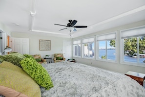 A bedroom with a large bed, a ceiling fan, a view of the water, and a chair by the window.