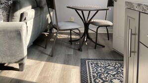 Ein kompakter Wohnbereich mit grauem Sofa, einem Esstisch mit zwei Stühlen, einer Küchenzeile und einem gemusterten Teppich.