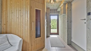 Un couloir intérieur en bois, avec un canapé blanc, une porte et une vue sur la verdure à l’extérieur.