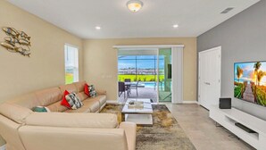 Una sala moderna con un sofá color beige, una mesa de centro y un televisor de pantalla plana. Hay una puerta corrediza de vidrio que da a un patio exterior con piscina.