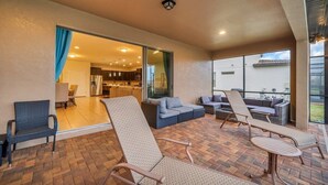 A covered patio with wicker furniture, a kitchen visible through an open door, and a large window offering a view of the outdoors.