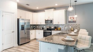 Cocina moderna con armarios blancos, electrodomésticos de acero inoxidable, encimeras de granito y una isla central con taburetes de bar.